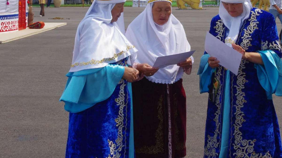 Three Kazakh women in traditional kimeshek headdress and blue velvet national costumes at a cultural event